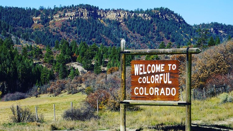 A Welcome To Colorado sign in the foreground -- brow with white text. In the background there are rolling hills of varying sizes with a mix of terrain -- dry grass to stone. Deep green pines cover the landscape as well.